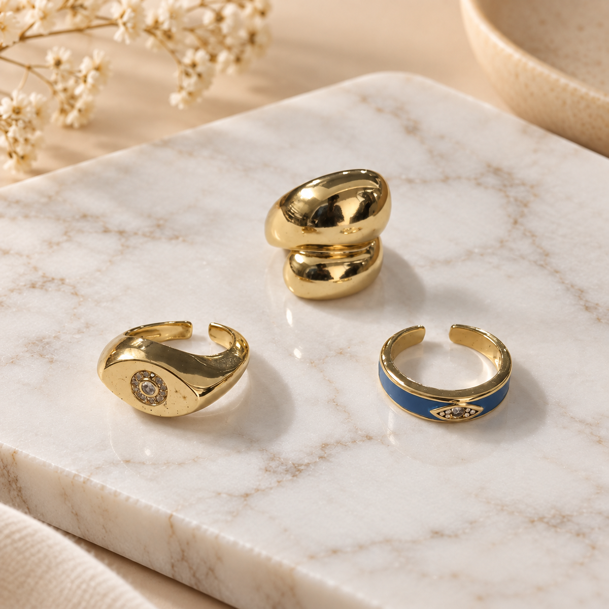 Three gold-tone rings arranged on a white marble surface, featuring smooth polished designs and subtle blue and crystal details, with soft floral accents in the background.