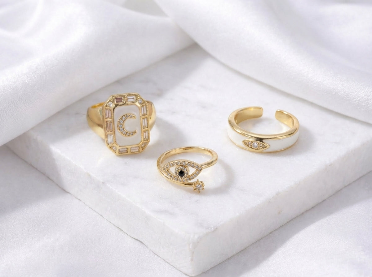 Gold-tone ring stack with ivory enamel band, blue moon ring, and evil eye rings with crystal detailing