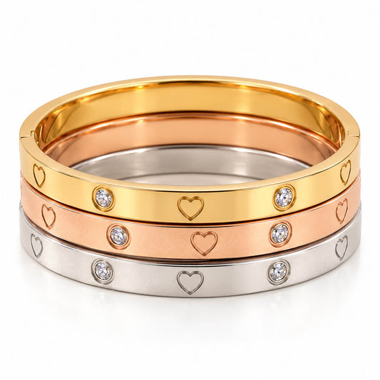 A product photo of a stacked set of mixed-metal bangles in gold, rose gold, and silver-tone finishes, featuring engraved heart outlines and bezel-set crystal stones.