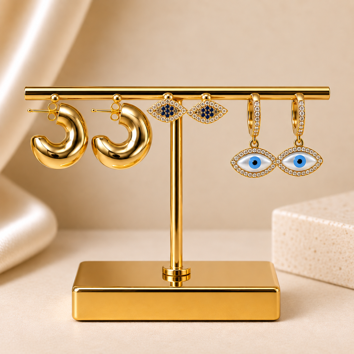 Gold-tone earring set displayed on a gold stand, featuring chunky hoops, blue Evil Eye studs, and crystal Evil Eye drop earrings.