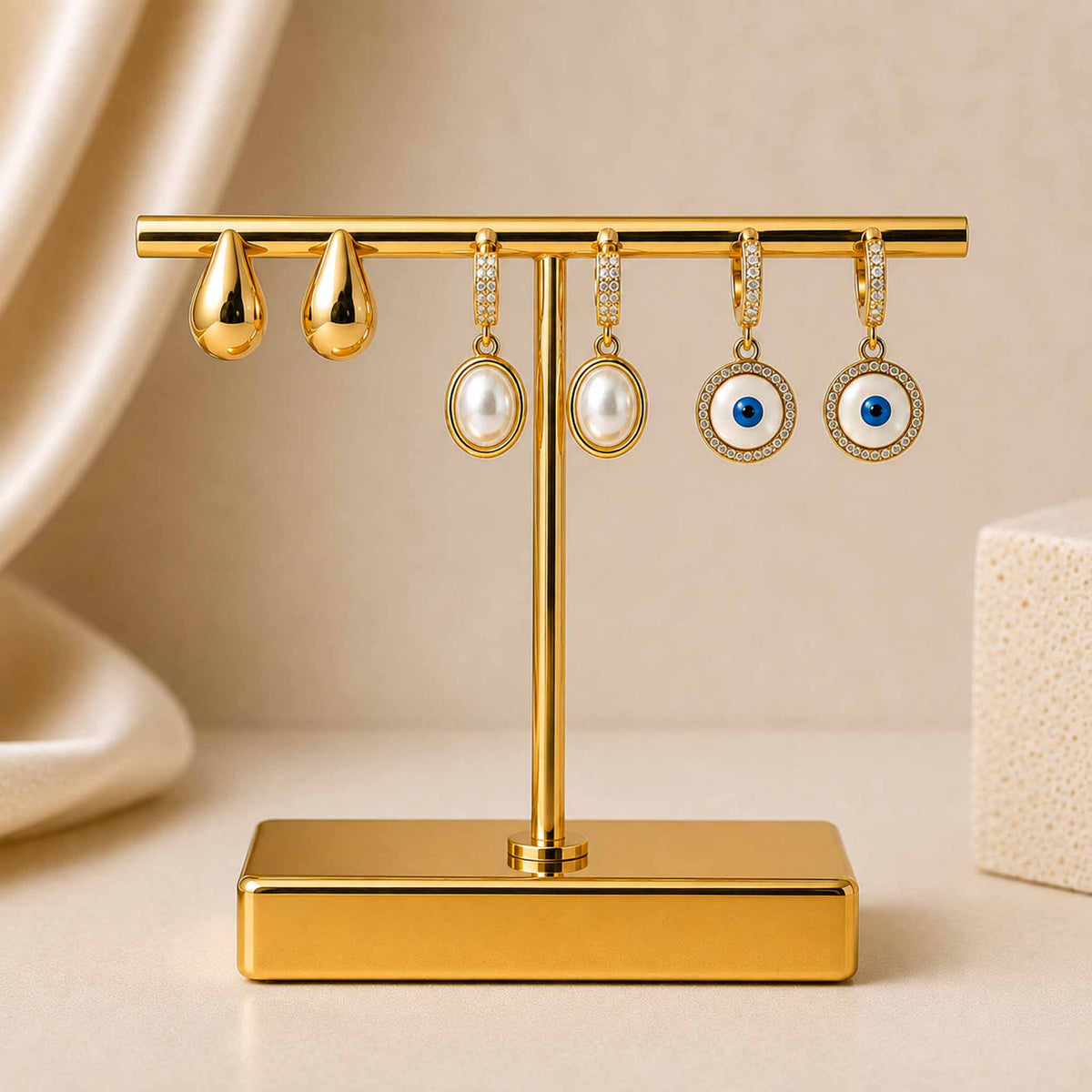 Gold-tone earring stack featuring polished teardrop stud earrings, oval pearl dangler earrings with crystal-studded tops, and circular blue evil eye dangler earrings, displayed on a gold jewellery stand with a neutral beige background.