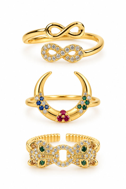 Three gold statement rings featuring an infinity duo ring, a colorful crescent crystal ring, and a bold butterfly-inspired pavé ring with green gemstone accents on a clean white background.