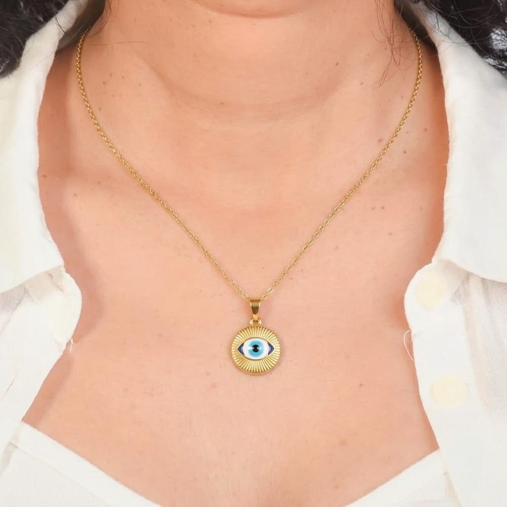Close-up view of a model wearing a fine gold chain necklace featuring a round gold pendant with radiating sunburst textures and an almond-shaped blue Evil Eye in the center. The pendant rests on the chest framed by an open white collared shirt, set against a dark gray border.