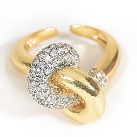 The image shows a gold-toned open cuff ring featuring an interlocking knot design. One loop is polished gold, while the other is densely embellished with clear pavé-style stones, creating a bold, luxurious contrast. It is photographed against a clean white background.