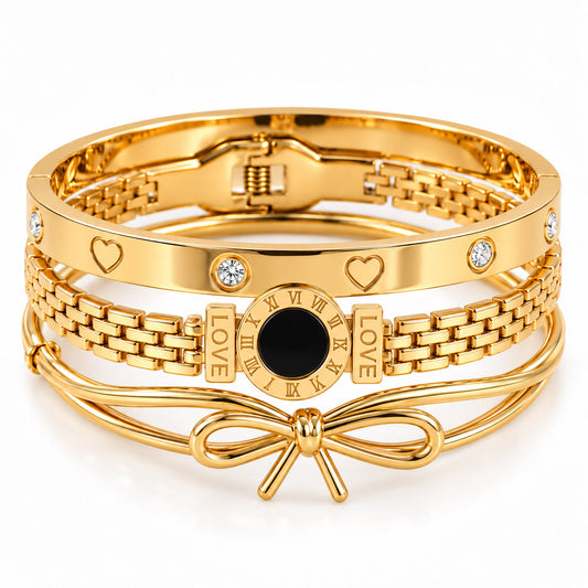 A product photo of a stacked set of polished gold-tone bracelets featuring an interlocking chain band, a bangle with engraved hearts and crystal stones, a link chain with roman numeral "LOVE" charms and a black disk, and a smooth wire bracelet featuring a sculptural bow.