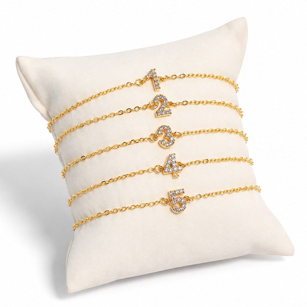 Gold chain lucky number crystal bracelets displaying numbers 1 through 5 on a cream jewelry display pillow.
