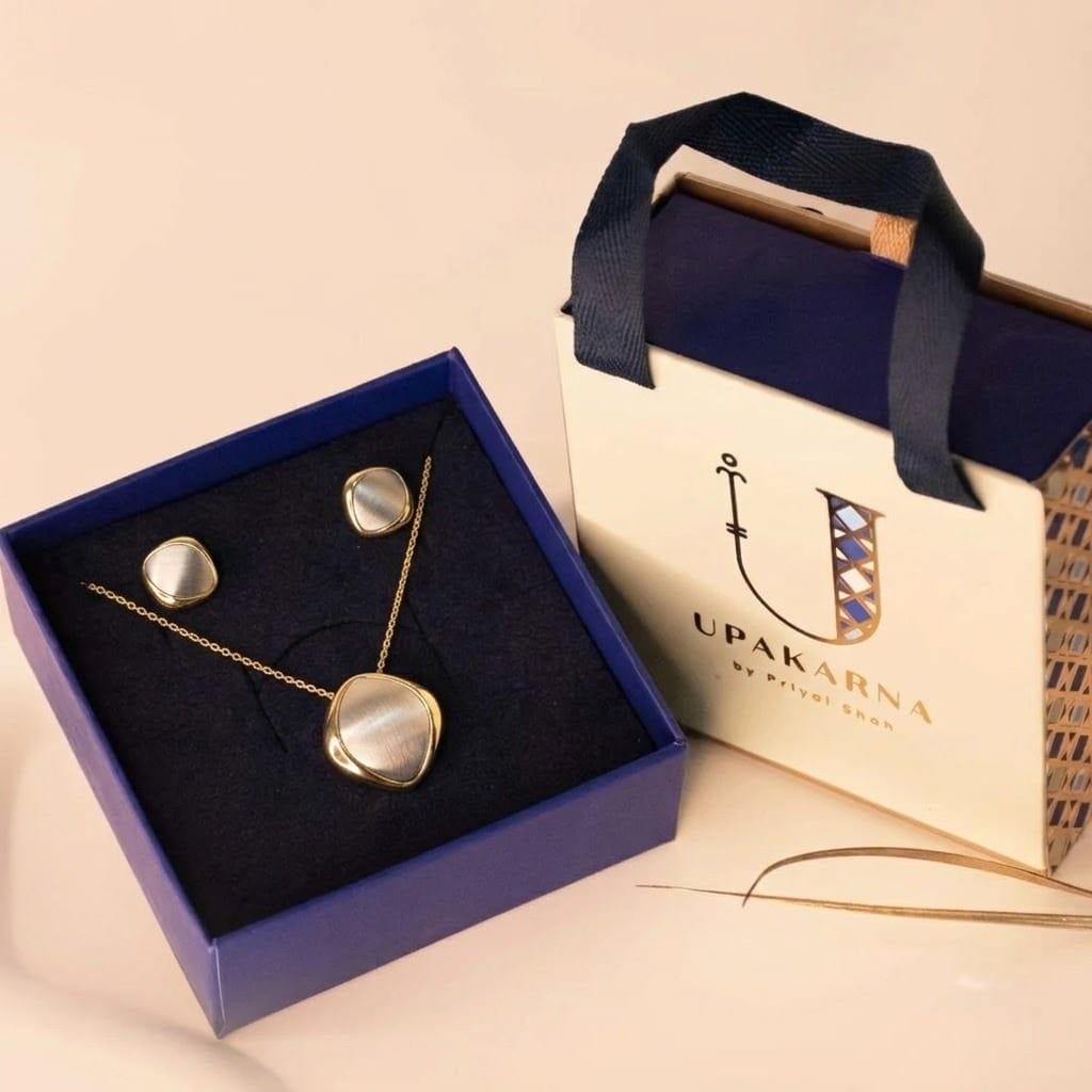An elegant jewelry set presented inside a dark blue display box next to a cream "UPAKARNA by Priyal Shah" gift bag. Displayed on the navy velvet interior, the set features a fine gold-chain necklace with an organic, wavy-edged cushion pendant showcasing a brushed silver central stone, paired with matching square cushion stud earrings.