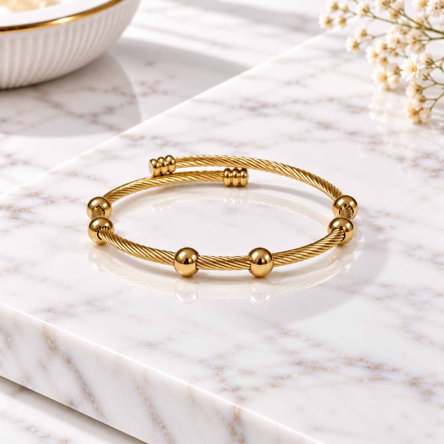 Gold twisted cable cuff bangle with polished bead detailing, displayed on a marble surface.