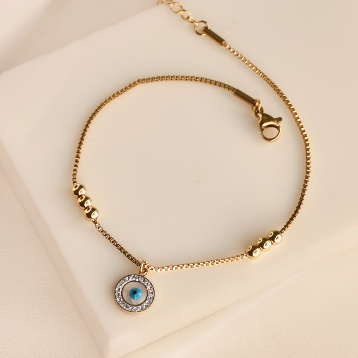 Delicate gold-tone anklet featuring a round blue evil eye charm surrounded by sparkling clear stones, with a fine chain and polished gold bead accents, arranged on a cream display surface against a soft beige background.
