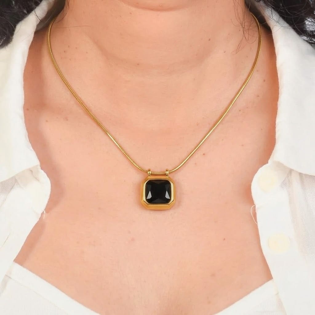 Close-up view of a model wearing a gold snake chain necklace holding a square-cut black gemstone pendant with a smooth gold bezel setting. The necklace rests on the chest framed by an open white collared shirt, set against a dark gray background frame.