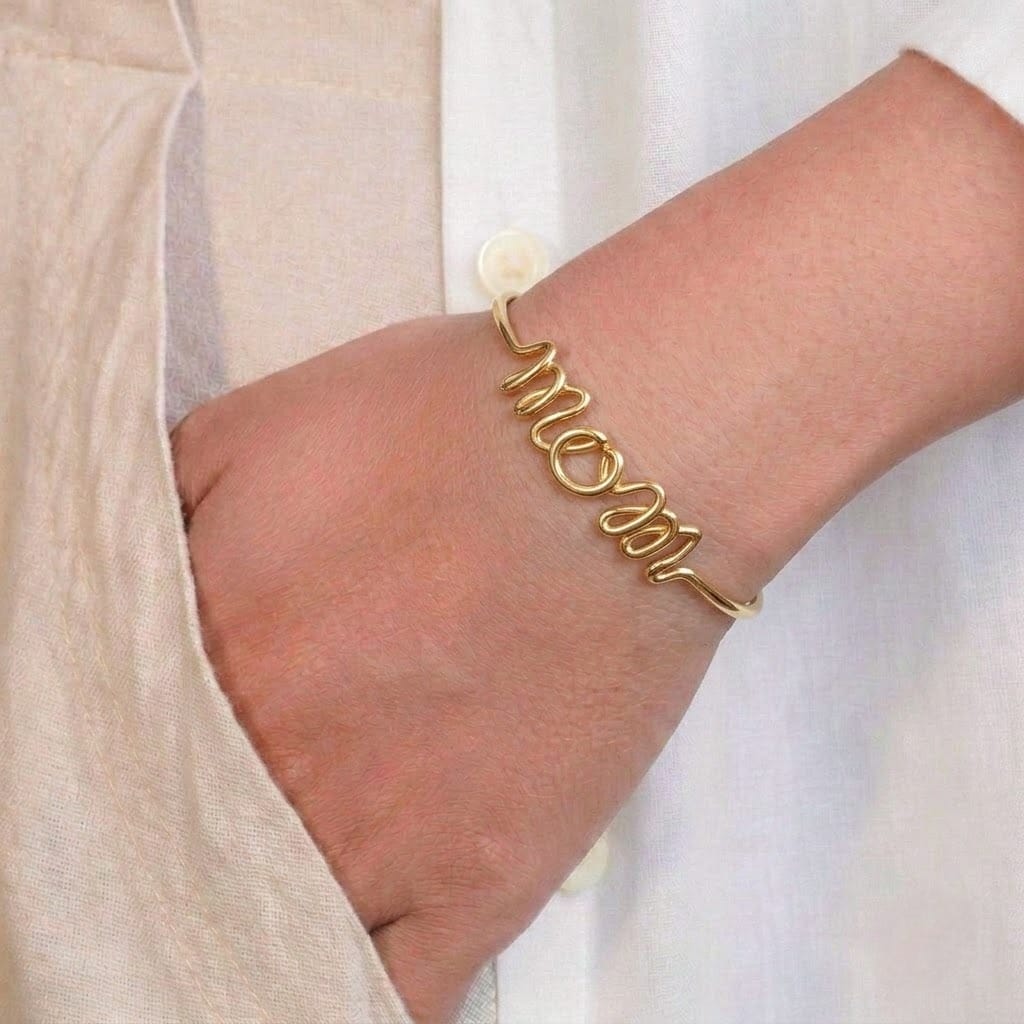 A close-up of a person’s wrist wearing a minimal gold-toned bangle designed in a cursive script forming the word “mom.” The sleek and sentimental bracelet is styled against a soft beige outfit, creating an elegant and personal look.