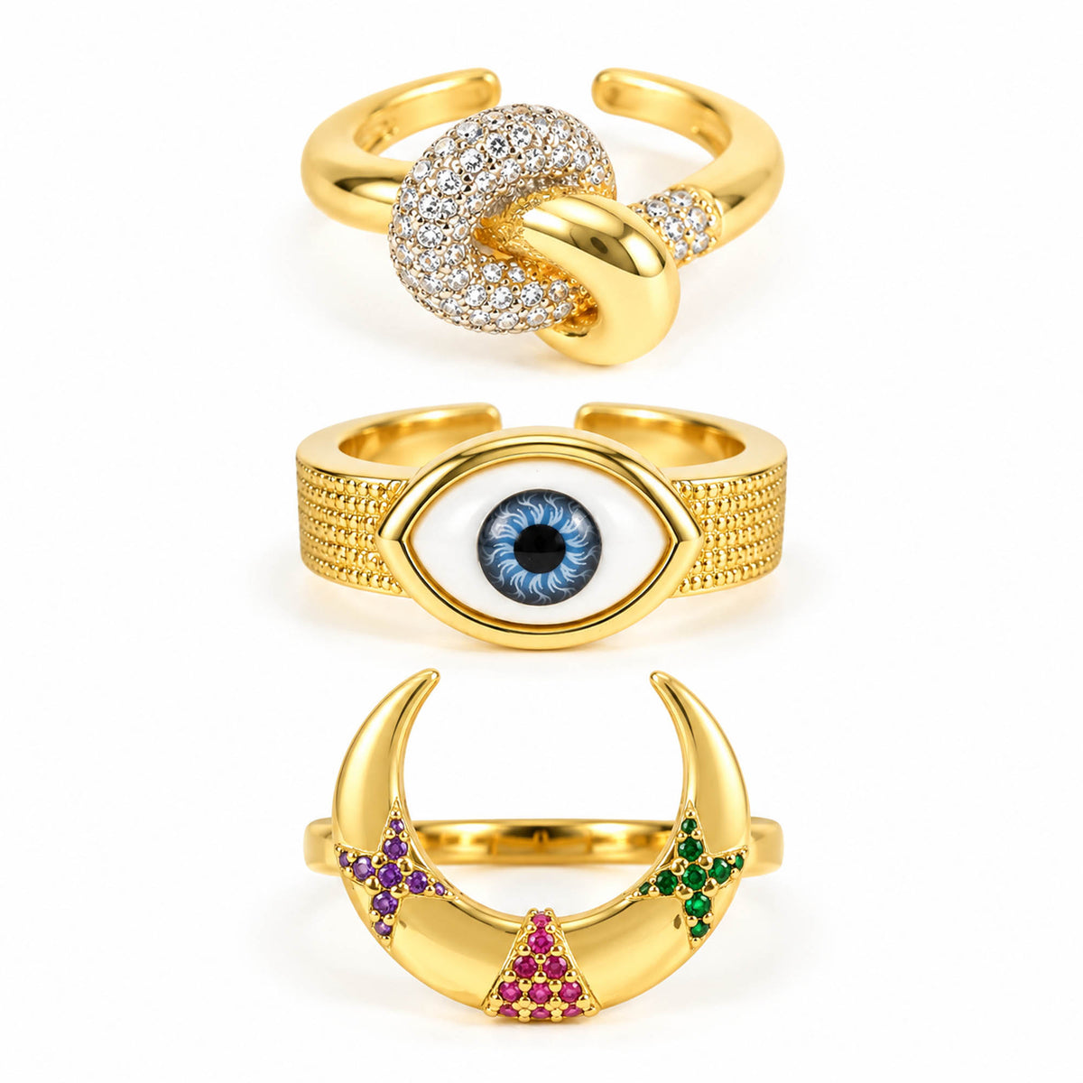 A product photo of three gold-tone adjustable rings featuring an intertwined knot band encrusted with crystals, a textured band centered with an enamel evil eye motif, and a crescent moon ring embellished with purple, pink, and green crystal clusters.