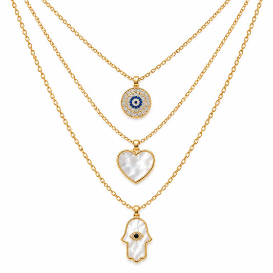 A product photo of a layered gold-tone necklace set featuring a circular evil eye pendant surrounded by rhinestones, a heart-shaped white mother-of-pearl pendant, and a white mother-of-pearl Hamsa hand pendant with a crystal-accented center.
