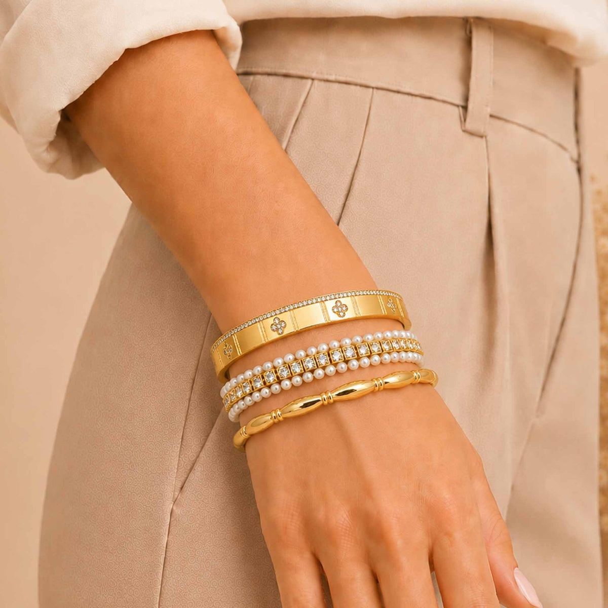 Gold bangle stack worn on a wrist, featuring a wide embellished cuff, pearl and crystal bracelet, and polished gold bangle with a beige outfit.