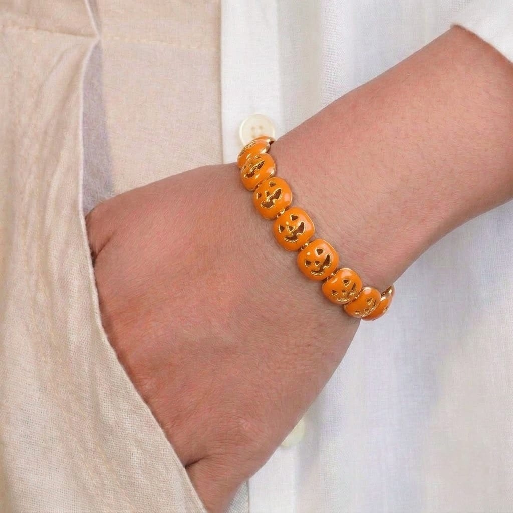A close-up of a person’s wrist wearing a playful orange beaded bracelet made of small pumpkin-shaped charms with carved jack-o’-lantern faces. The bracelet adds a fun, festive touch and is styled against a soft beige outfit.