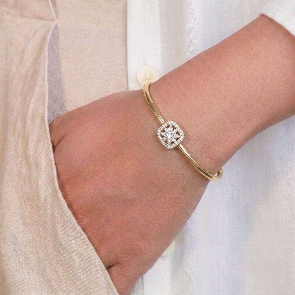 A close-up of a person’s wrist wearing a minimal gold-toned bangle featuring a small square centerpiece encrusted with tiny sparkling stones in a geometric pattern. The delicate bracelet is styled against a soft beige outfit, creating an elegant and refined look.