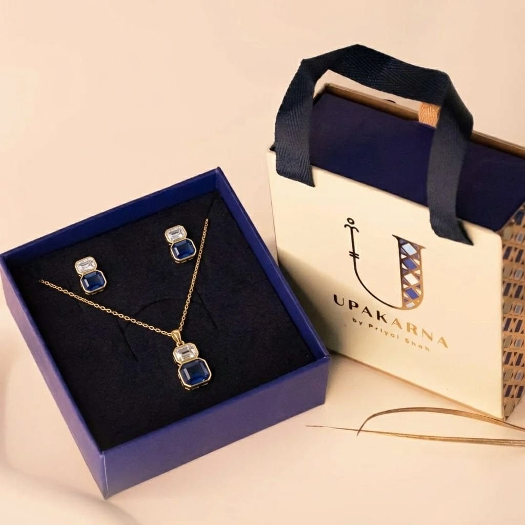 A gold jewelry set presented inside a dark blue display box beside a cream "UPAKARNA by Priyal Shah" gift bag. Arranged on the navy velvet interior, the collection features a fine-chain necklace showcasing a two-stone drop pendant with a clear emerald-cut crystal stacked above a larger sapphire-blue gemstone, accompanied by a pair of matching stacked-stone stud earrings.