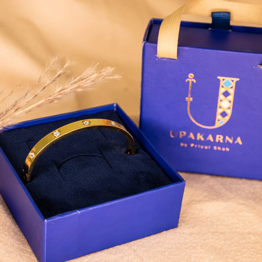 Gold anti-tarnish bangle featuring delicate heart motifs and sparkling crystal accents, elegantly displayed in an open navy blue Upakarna jewellery box with a matching branded gift bag. Warm beige satin styling, golden lighting, and a premium luxury gifting aesthetic.