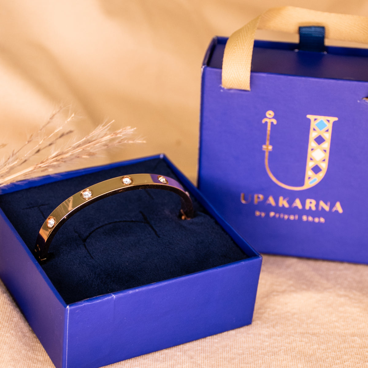 RoseGold anti-tarnish bangle featuring a sleek polished finish and evenly spaced sparkling crystal accents, presented in an open navy blue Upakarna jewellery box with a matching branded gift bag. Styled against a warm beige background with delicate dried botanicals and soft golden lighting for a premium gifting aesthetic.