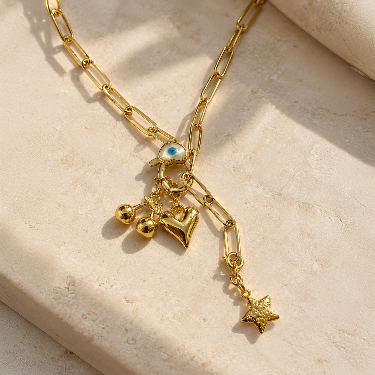 Gold-plated paperclip chain necklace featuring a blue Evil Eye centerpiece with dangling gold cherries, heart, and textured star charms, styled on a beige stone surface with warm natural sunlight and soft shadows.