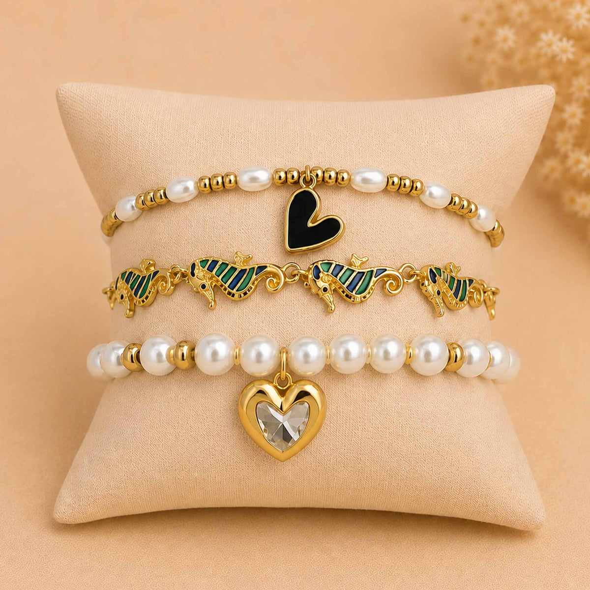 Golden pearl bracelet stack displayed on a cream cushion, featuring a black heart pearl bracelet, green seahorse enamel bracelet, and pearl heart charm bracelet.