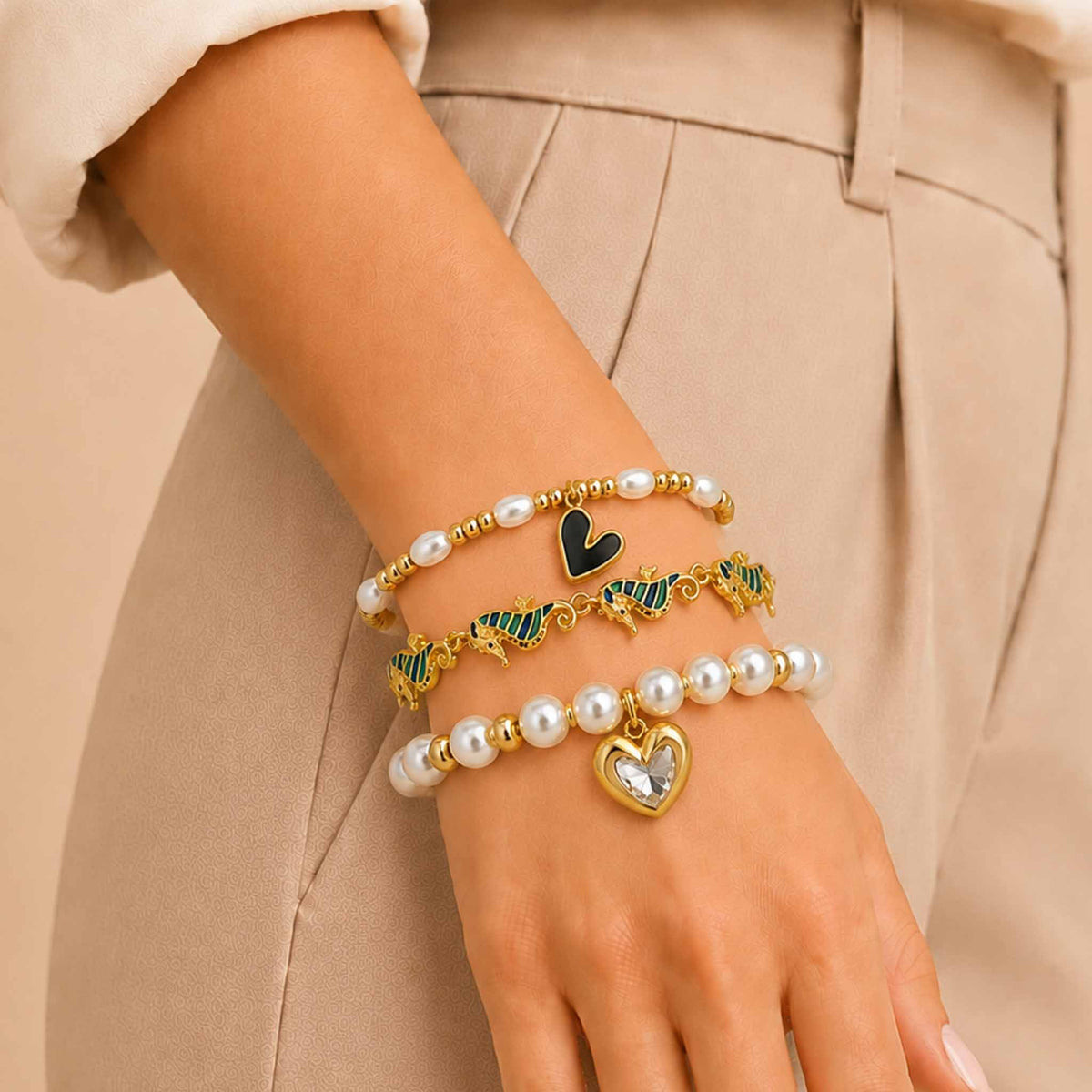 Golden pearl bracelet stack worn on a wrist, featuring a black heart pearl bracelet, green seahorse enamel bracelet, and pearl bracelet with a crystal heart charm.