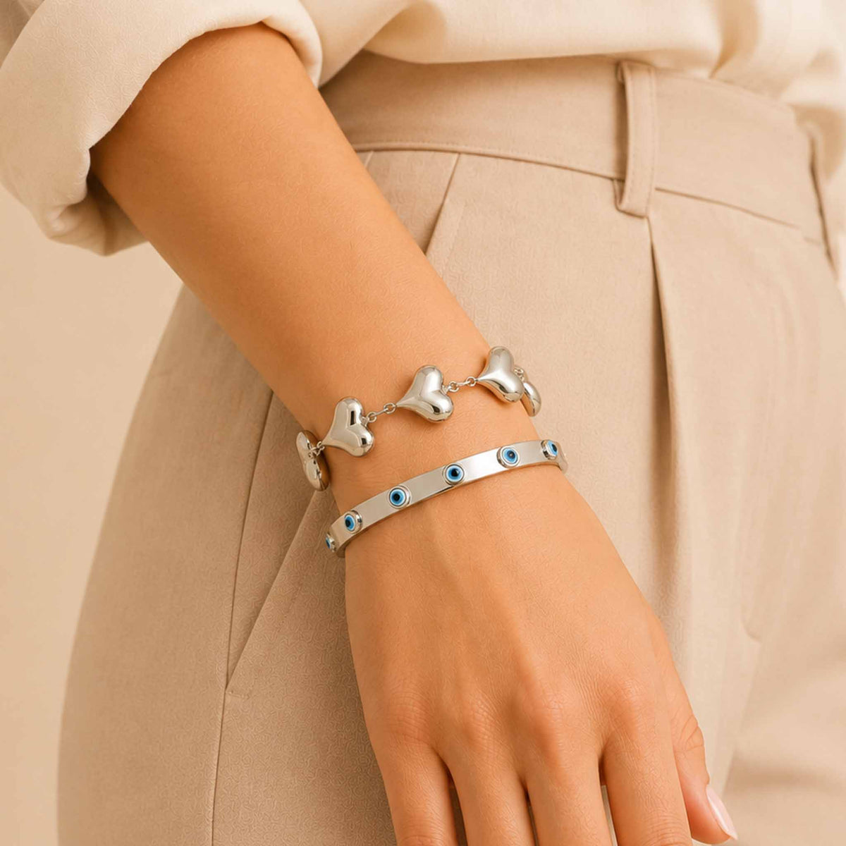 Silver bracelet stack worn on a woman’s wrist, featuring a chunky heart-link bracelet and an enamel Evil Eye bangle.