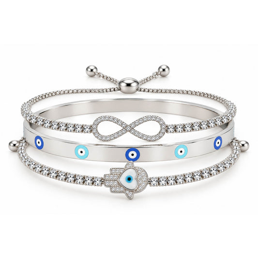 A product photo of a stacked set of silver-tone bracelets featuring an adjustable crystal band with an infinity symbol, a solid bangle with blue-and-white evil eye motifs, and a crystal tennis bracelet centered with a Hamsa hand charm containing a heart-shaped evil eye.