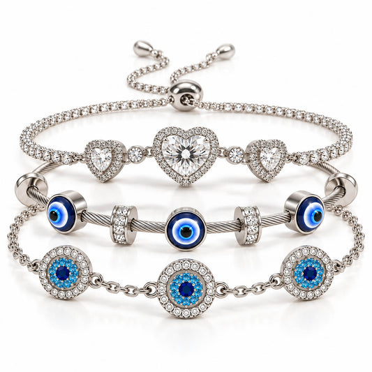A product photo of a stacked set of silver-tone bracelets featuring an adjustable crystal tennis chain with heart-shaped pendants, a twisted cable wire band with blue-and-white evil eye charms, and a link chain with paved circular evil eye stations.