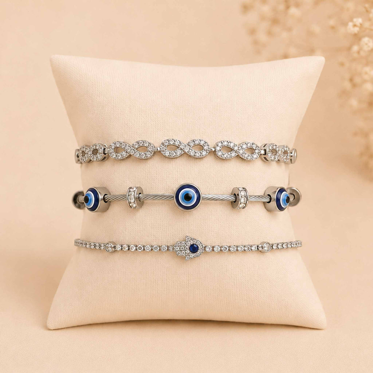 Silver bracelet stack displayed on a cream cushion, featuring an infinity crystal bracelet, Evil Eye bangle, and sparkling blue Evil Eye tennis bracelet.
