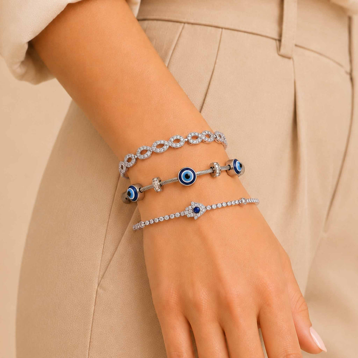 Silver bracelet stack worn on a wrist, featuring an infinity crystal bracelet, Evil Eye bangle, and sparkling blue Evil Eye tennis bracelet with a cream outfit.