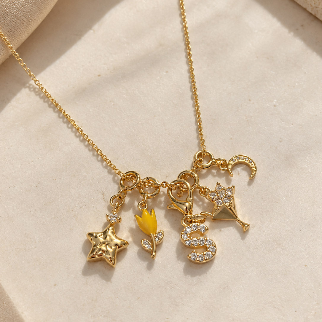 Gold-plated charm necklace featuring a star, yellow tulip, sparkling “S” initial, floral champagne glass, and crescent moon charms on a delicate chain, displayed on a clean white background.