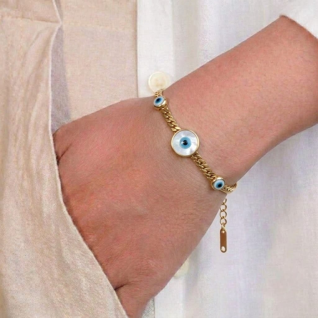 Gold-tone braided Evil Eye bracelet worn around a person’s wrist, featuring three blue and white Evil Eye charms with black centers. The bracelet has a textured woven design, adjustable extension chain, and gold-tone clasp. The image is styled with a light beige outfit against a soft white fabric background.