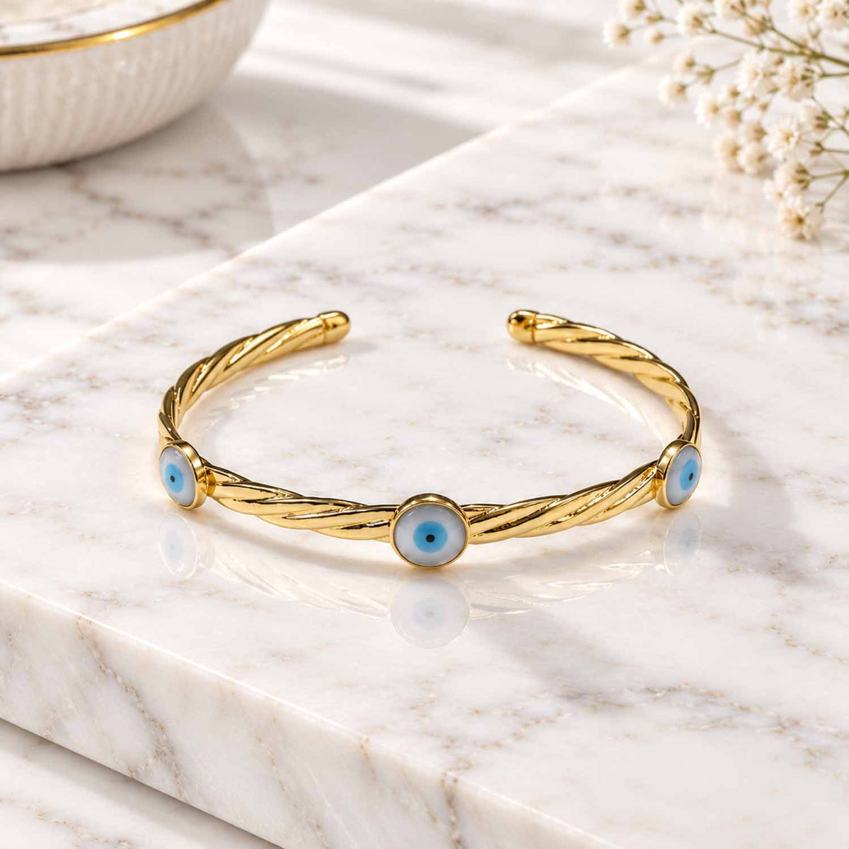 Gold twisted evil eye cuff bangle with three blue evil eye motifs, displayed on a marble surface with soft natural lighting.
