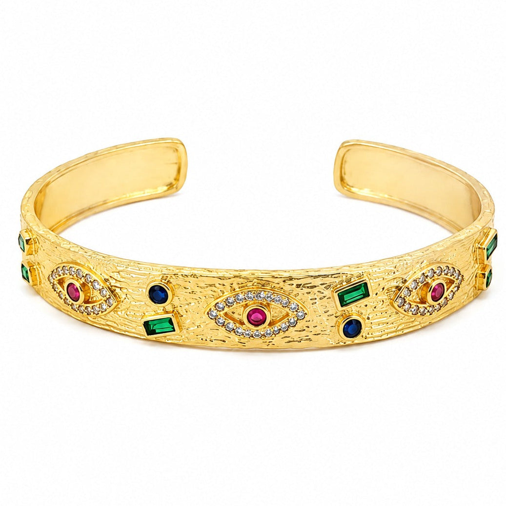 Anti-tarnish textured gold open cuff bracelet featuring evil eye motifs with pavé crystal borders and ruby-red centers, interspersed with bezel-set green emerald baguettes and deep blue sapphire round stones.