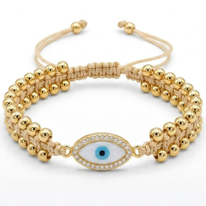 Gold-tone evil eye bracelet with polished round beads, a beige braided cord, and a blue-and-black evil eye charm bordered with sparkling clear crystals, displayed on a white background.