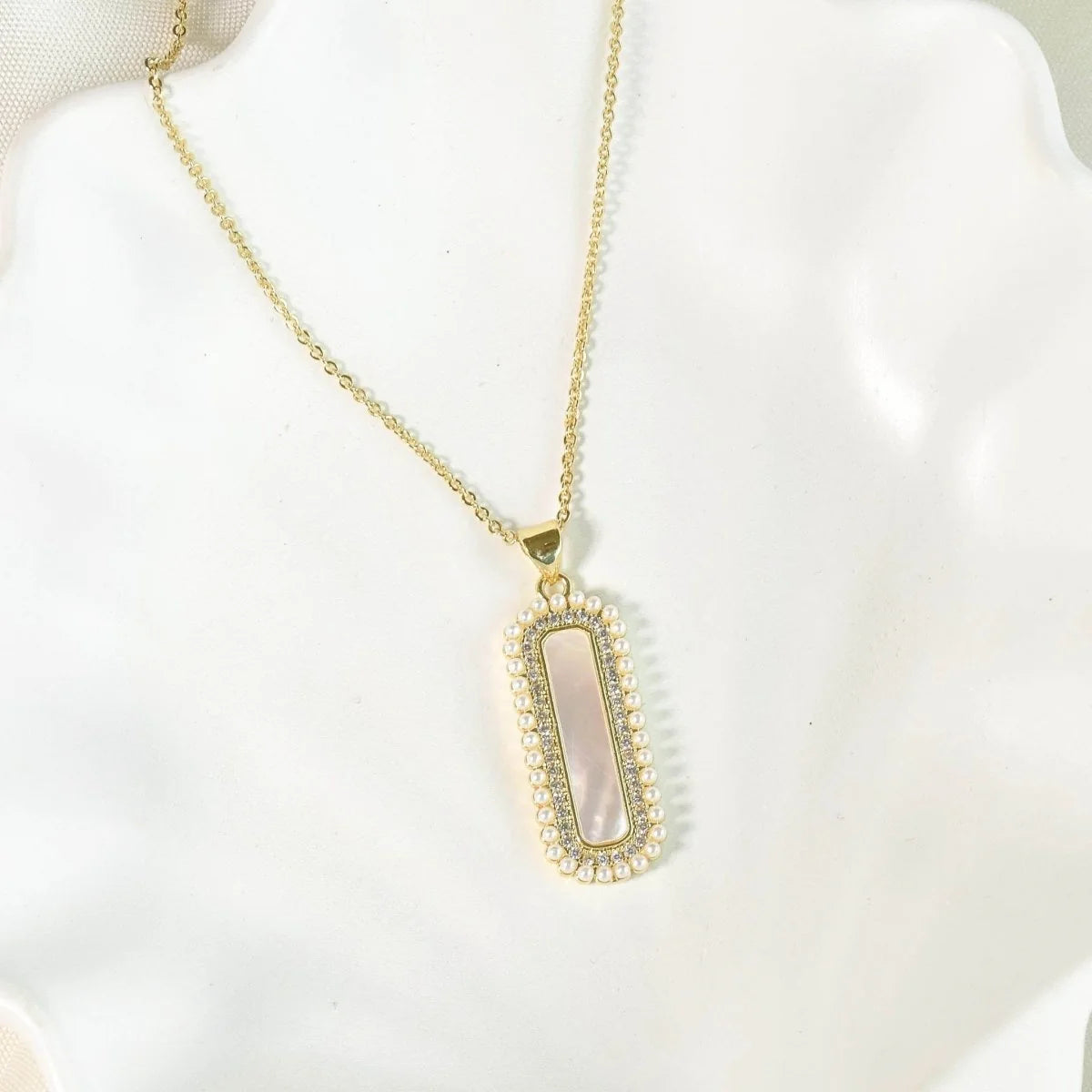 The image shows a delicate gold-toned necklace displayed on a white mannequin bust. It features a slim chain and an elongated rectangular pendant framed with small white pearls and a pavé-style crystal border, creating a refined, elegant look.