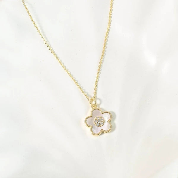 The image shows a delicate gold-toned necklace with a flower-shaped pendant. The pendant has a smooth gold outline, a white/mother-of-pearl-like center, and a small cluster of clear sparkling stones at the center. It is photographed against a clean white background.