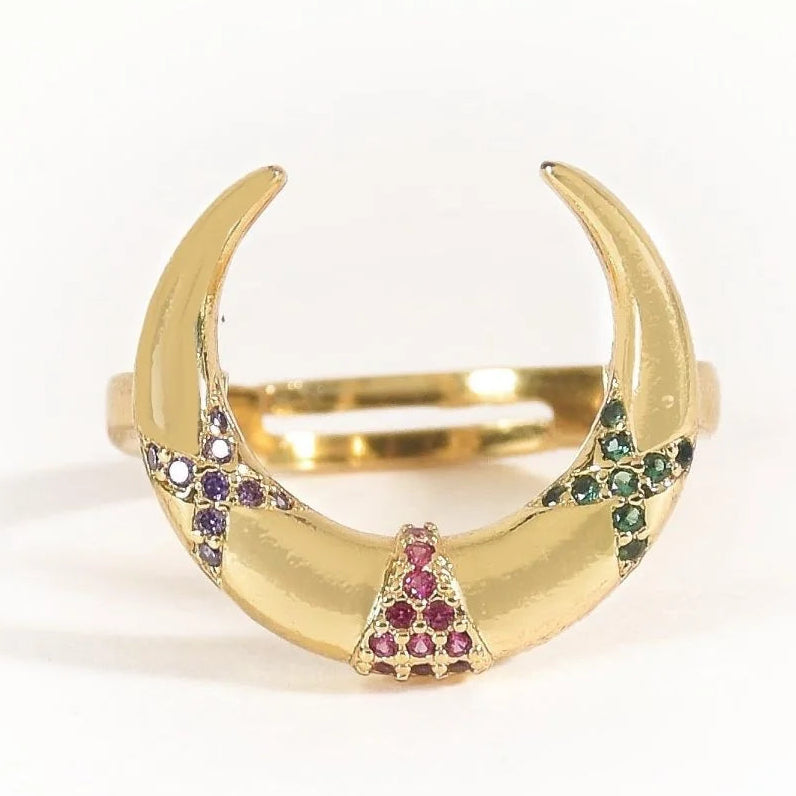 The image shows a gold-toned open cuff ring with a bold curved, horseshoe-like design. It features colorful pavé-style gemstone accents in pink, purple, green, and clear tones arranged across the front, creating a vibrant statement look. The ring is photographed against a clean white background.