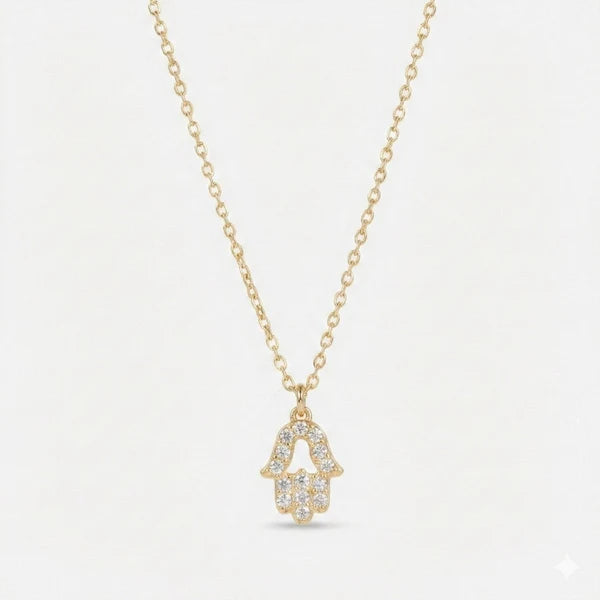 The image shows a delicate gold-toned necklace with a Hamsa hand-shaped pendant. The pendant is fully embellished with clear pavé-style stones and has a small teardrop-shaped stone at the bottom. It is photographed against a clean white background.