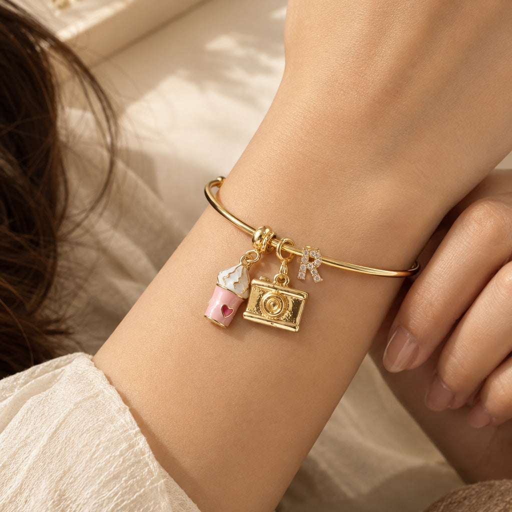 Gold open cuff bangle worn on a wrist, featuring a sparkling letter R charm with a pink cupcake and camera charms.