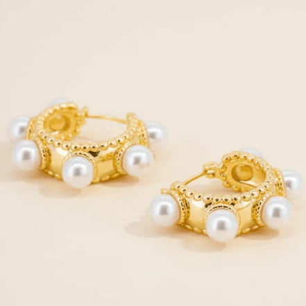 Gold-tone huggie hoop earrings decorated with multiple white pearl accents and delicate beaded detailing, photographed on a warm cream background.