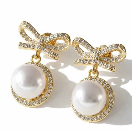 Gold-tone bow drop earrings featuring large white pearl-like stones surrounded by clear crystals, with crystal-studded bow-shaped tops.