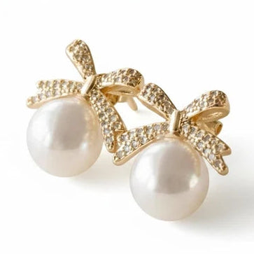 Gold-tone pearl stud earrings with large round white pearls and pavé crystal bow-shaped accents, displayed on a clean white background.