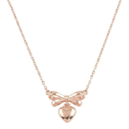 The image shows a delicate rose-gold necklace with a fine chain, featuring a detailed bow-shaped pendant with layered ribbon loops and a small polished heart charm hanging from the center. The jewelry is photographed against a clean white background.