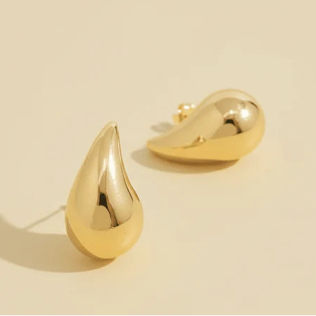 Gold-tone teardrop stud earrings with a smooth, polished sculptural finish, photographed on a warm beige background.