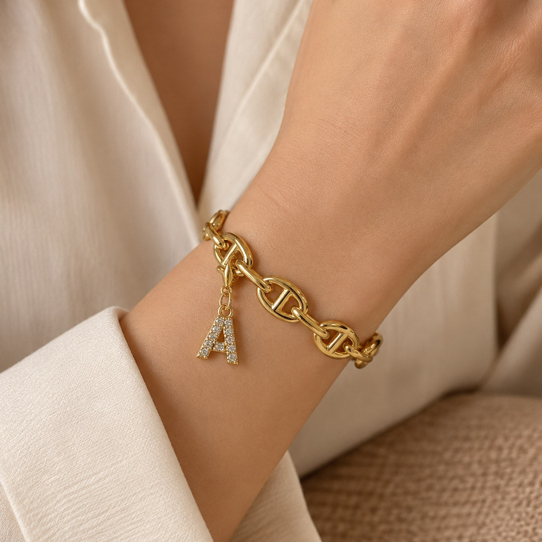 Close-up of a person wearing a gold-tone statement bracelet with linked oval chain detailing and a dangling crystal-studded letter “A” charm, styled with a white outfit against a warm neutral background.