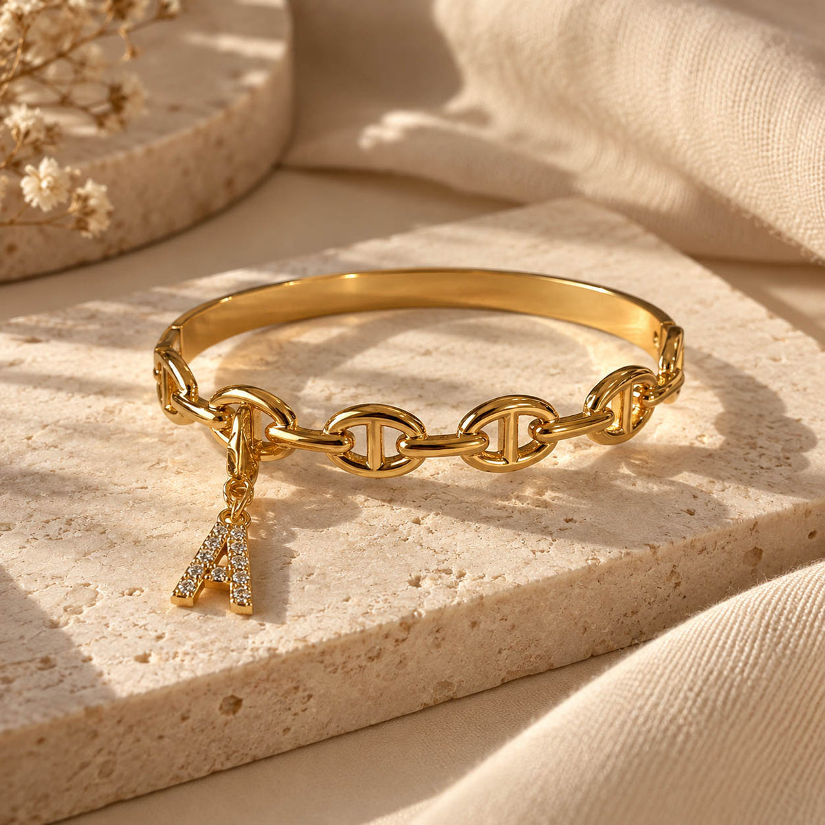 Gold-tone statement bracelet with a smooth cuff band, linked oval chain detailing, and a dangling crystal-studded letter “A” charm, styled on a beige stone surface with warm neutral fabric and soft natural lighting.