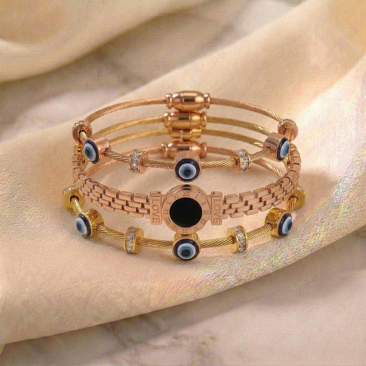 A studio product photograph of a curated stack of rose gold-toned charm bracelets resting on a soft, draped neutral fabric background. The stack features cable-wire bands accented with dark blue and white evil eye beads and sparkling crystal station spacers, centered with a distinctive watch-link band featuring an engraved Roman numeral dial and "LOVE" inscriptions flanking a central black stone.   