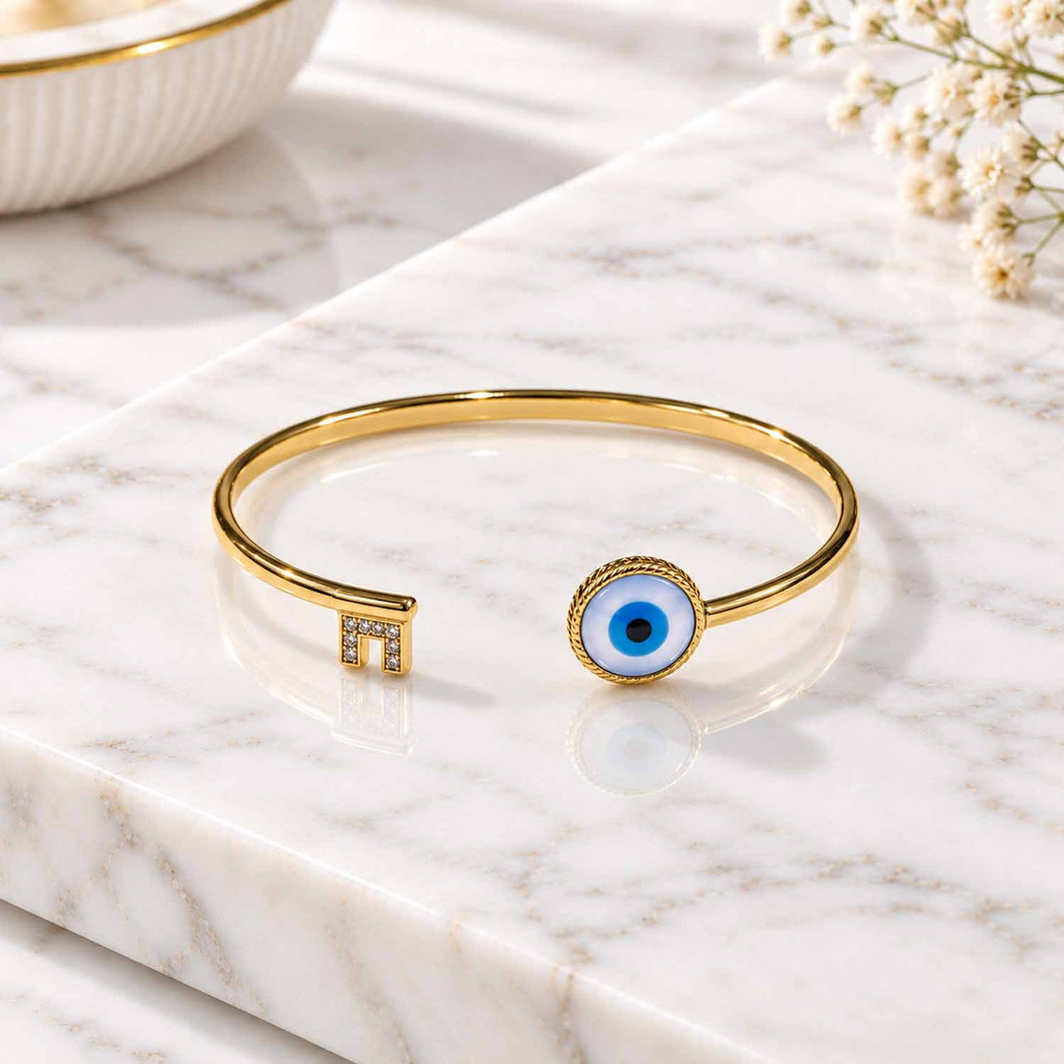 Gold open cuff bangle featuring a blue evil eye centerpiece and a sparkling stone-studded geometric end detail, displayed on a marble surface with soft natural lighting.